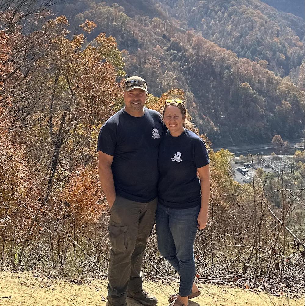 Two people are standing close together, smiling, in front of a scenic, forested mountain landscape on a sunny day. Both are wearing matching black T-shirts.