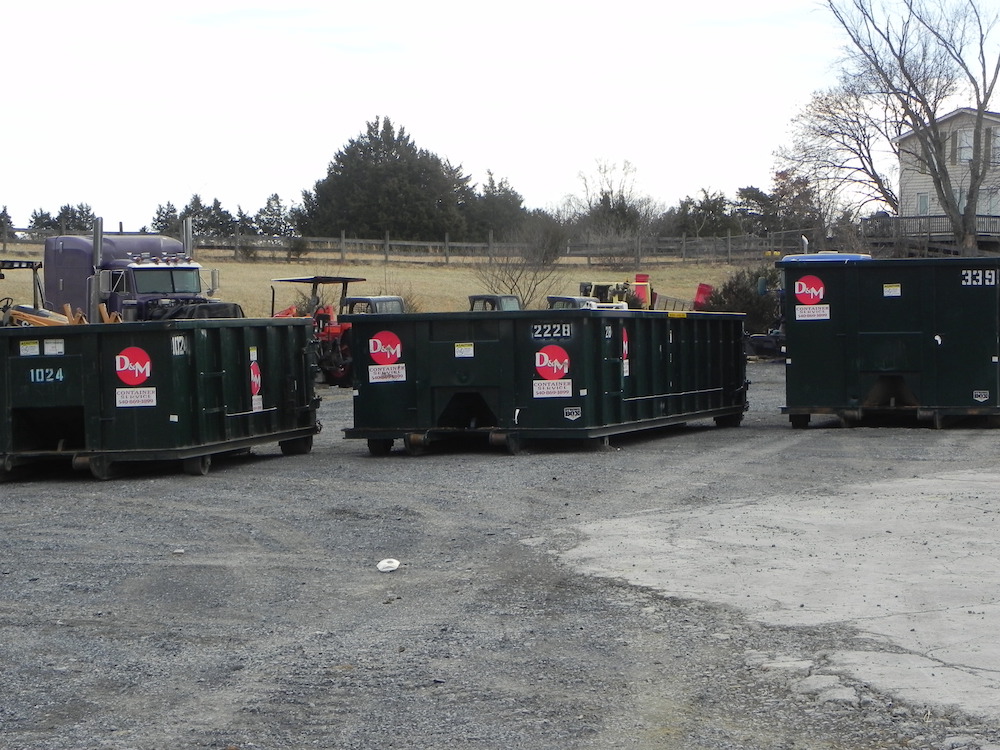 The image shows a collection of green dumpsters labeled "D&W" lined up in an outdoor gravelly area, indicating a waste management or construction site.