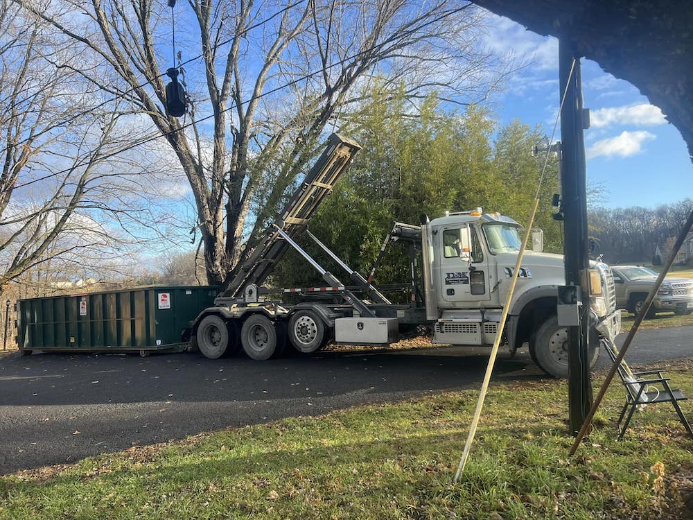 A large white roll-off truck is lifting a green dumpster under a clear sky. Trees are nearby, and it's parked on a gravel area beside a road.