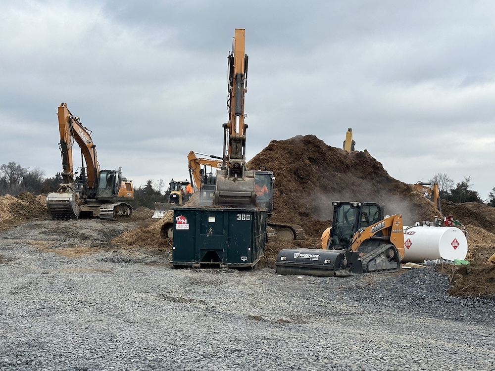 A construction site with various heavy machinery including excavators and a skid steer loader. There's a large dirt pile and overcast skies above.