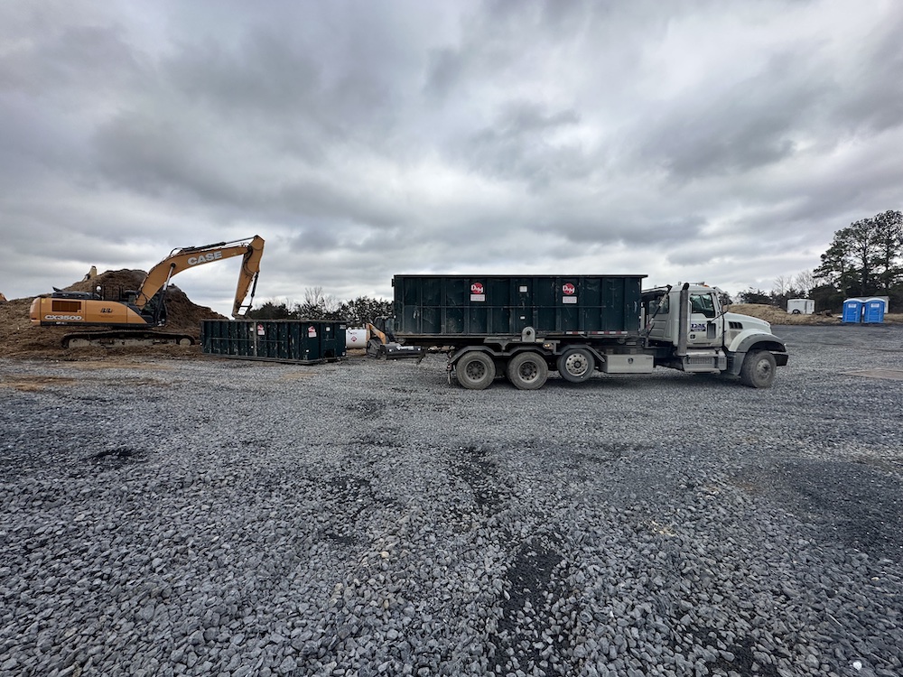 A construction site with an excavator, a parked truck with a dumpster trailer, a portable toilet, and an overcast sky. The ground is covered with gravel.