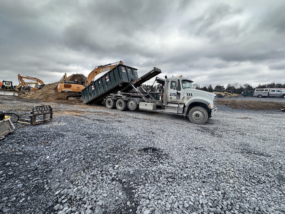 A dump truck is tilted to unload its contents at a construction site, with excavators in the background under a cloudy sky.