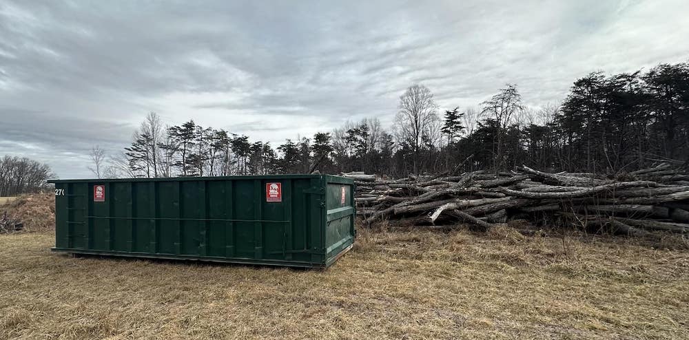 A large green dumpster sits on a grassy field beside a pile of logs, under a cloudy sky with trees in the background.